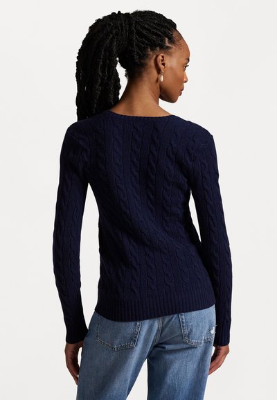 Tamsiai mėlynas megztinis su pynimo raštu, ilgomis rankovėmis ir apvaliu iškirpimu. Pora šviesiai mėlynų džinsų su nusidėvėjimo detalėmis.