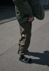 Veste matelassée verte, pantalon cargo olive avec poches et étiquette Carhartt, sandales noires avec rayures grises, portées avec des chaussettes noires.