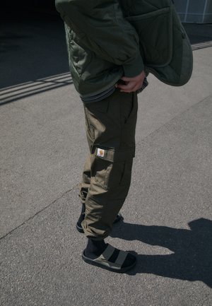 Chaqueta acolchada verde, pantalones cargo oliva con bolsillos y etiqueta de Carhartt, sandalias negras con rayas grises, usadas con calcetines negros.