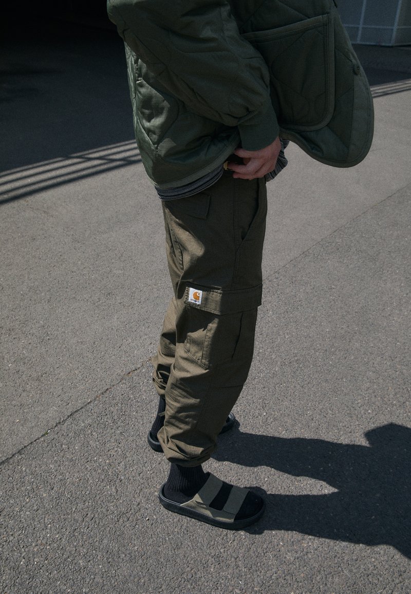 Veste matelassée verte, pantalon cargo olive avec poches et étiquette Carhartt, sandales noires avec rayures grises, portées avec des chaussettes noires.