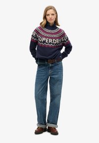 Maglione lavorato a maglia blu navy con motivo geometrico multicolore e logo sul petto; abbinato a jeans dritti blu chiaro e scarpe marroni.