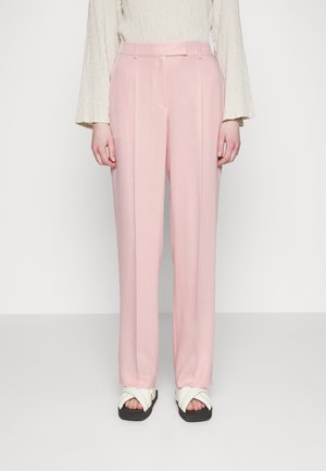 Pantalons roses clair avec une coupe ajustée, présentant une texture lisse et des détails de plis subtils ; associés à des sandales blanches à lanières croisées.