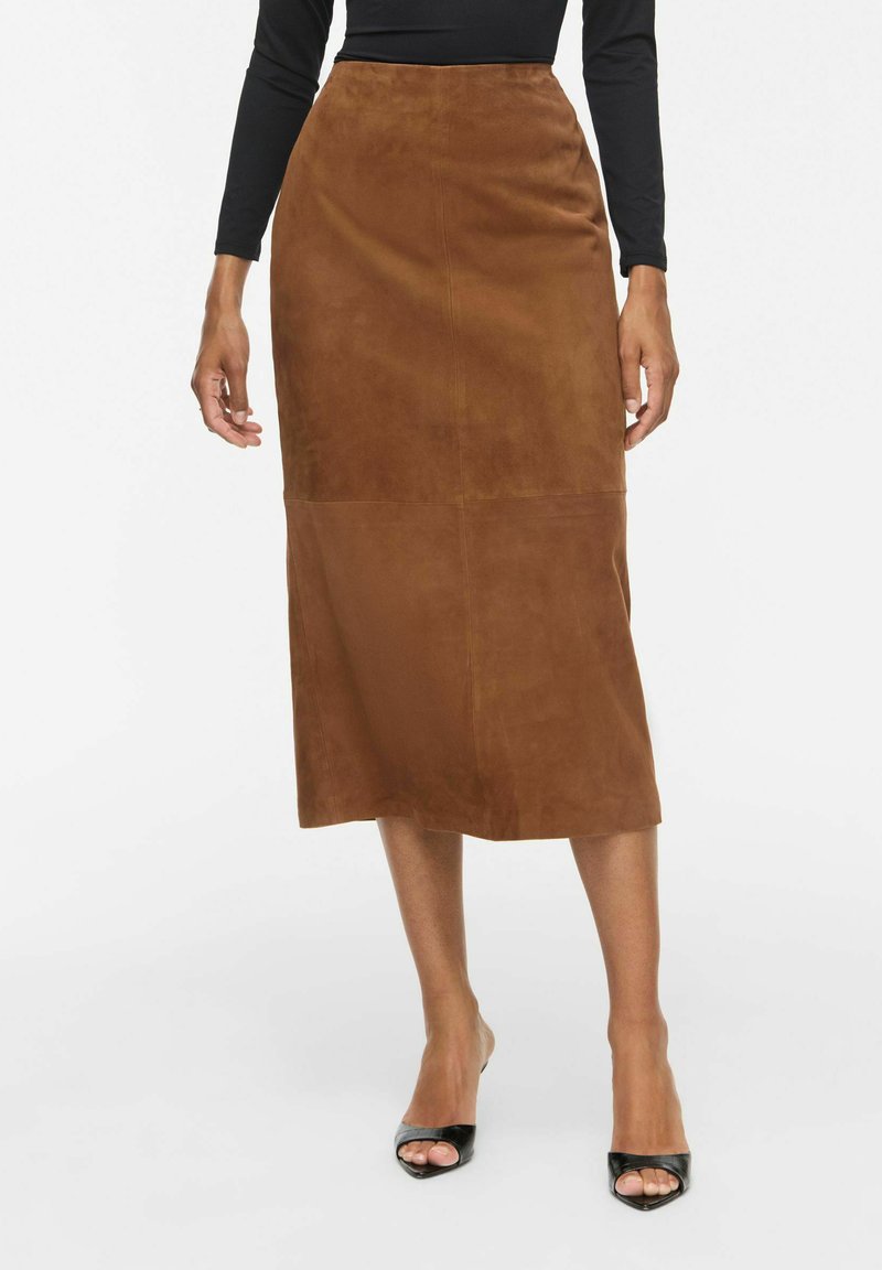 Personne portant une jupe midi en daim marron associée à des talons ouverts noirs et un haut noir à manches longues, debout sur un fond blanc.
