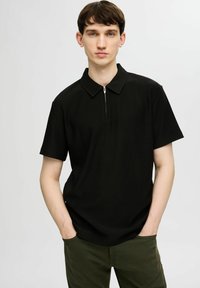 Czarny polo z prążkowanym wzorem, z kołnierzykiem na zamek, krótkimi rękawami i luźnym krojem, w zestawie z zielonymi spodniami. Prosty przód i minimalne detale.