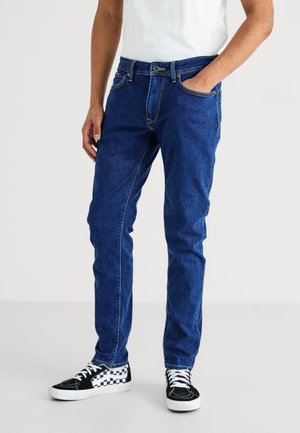 Homme portant un jean slim bleu, une main dans la poche et des baskets à carreaux noir et blanc sur un fond uni.