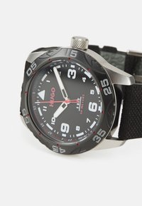 Reloj de pulsera negro y plateado con una correa de tela texturizada, esfera con números grandes, acentos rojos y bisel de acero inoxidable. Resistente al agua hasta 50 metros.