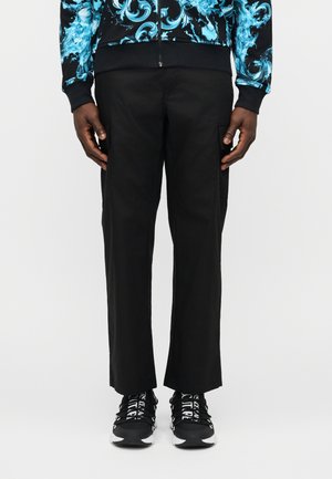 Man wearing black straight-leg pants, black and white sneakers with text on laces, and a blue patterned zip-up jacket.