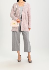 Cardigan rosa chiaro, texturizzato, aperto davanti; pantaloni grigi a vita alta; tacchi grigi; piccola borsa bianca con dettagli dorati; look morbido e stratificato.