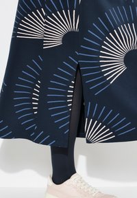 Marinblå kjol med ett geometriskt mönster av växlande blå och rosa linjer. Sidoflitsen avslöjar ett svart byxben och en ljusrosa sneaker.