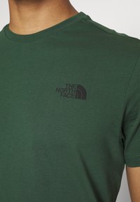 Grön bomullströja med korta ärmar och rundad halsringning, med en svart "The North Face"-logotyp tryckt på vänster bröst.