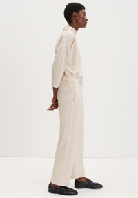 Beige longsleeve top met een ontspannen pasvorm en een omgeslagen kraag, gecombineerd met ruime, lichtbeige broeken en zwarte loafers.