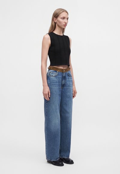 Jeune femme debout portant un crop top noir sans manches, un jean bleu taille haute à jambes larges avec une ceinture marron, et des chaussures noires.