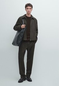 Giacca marrone con texture sopra un maglione scuro e pantaloni coordinati, abbinati a una borsa a tracolla in pelle nera e scarpe lucide nere.