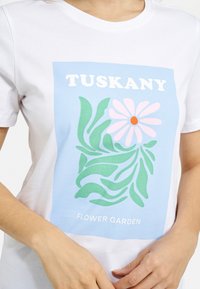 White cotton t-shirt with a blue graphic featuring the text "TUSKANY" and "FLOWER GARDEN," and floral and leaf designs in pink and green.