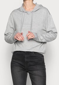 Osoba ubrana w jasnoszary hoodie z trokami i czarne dżinsy, stojąca z rękami lekko uniesionymi i palcami w luźnej pozycji.