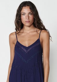 Robe bleu marine à fines bretelles, avec des découpes géométriques et des accents métalliques de couleurs variées sur le corps et l'ourlet. Texture de tissu douce.