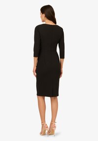 Schwarzes knielanges Kleid mit Dreiviertelärmeln, Nahtdetails, einem Reißverschluss auf der Rückseite und einer figurschmeichelnden Silhouette. Kombiniert mit hautfarbenen High Heels.