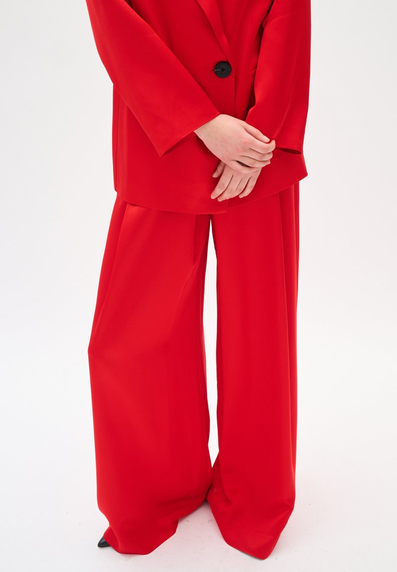 Blazer rojo con un solo botón negro, combinado con pantalones rojos de corte holgado. El material parece suave con un acabado mate.