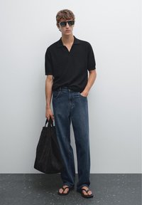 Uomo in polo nera e jeans blu sta in piedi con una borsa a tracolla nera, indossando occhiali da sole, di fronte a uno sfondo neutro. Posizione rilassata, stile casual.