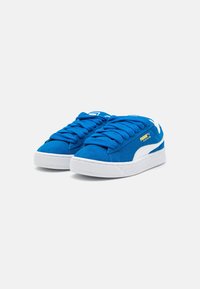 Zapatillas de ante azul con suelos de goma blanca, con cordones prominentes, una franja lateral blanca y un logo dorado en el costado.