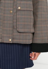 Giacca a scacchi nelle tonalità del beige, marrone e grigio, con bottoni a scatto dorati e polsino a costine nero. Indossata sopra una gonna a righe blu navy.