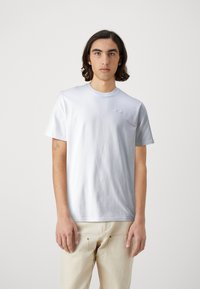Bílý bavlněný tričko s kulatým výstřihem, krátkými rukávy a nenápadným logem na hrudi. Model má na sobě světlé kalhoty.