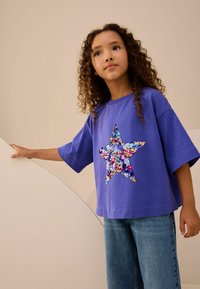 Camiseta de manga corta morada con un diseño de estrella de lentejuelas multicolores. Combinada con unos pantalones vaqueros de mezclilla azul claro, presentada sobre un fondo neutro.