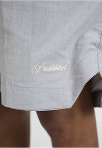 Shorts gris clair avec le mot brodé en blanc « Interdit » près de l'ourlet, porté par une personne à la peau foncée.
