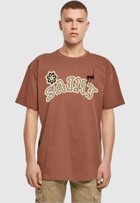 Brun oversized t-shirt med hvid og sort grafisk tekst "SAINT" og blomsterdesign. Lavet af blødt bomuld med en afslappet pasform.