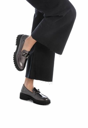Mocasines de cuero gris con un acabado texturizado, que presentan un acento decorativo de cadena y suelas de goma negras gruesas. Combinados con pantalones negros cortos.