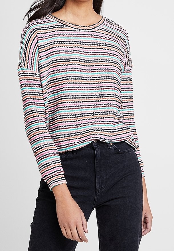Femme portant un haut à manches longues rayé aux couleurs pastel, rentré dans un jean noir taille haute, sur un fond uni.