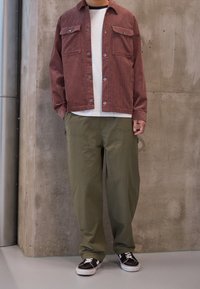 Rode corduroy overhemdjacket met voorzakken, witte t-shirt, groene lichte broek en zwarte sneakers met witte zolen. Glad oppervlak.