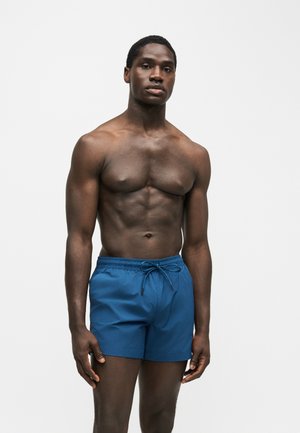 Homme musclé debout devant un fond blanc, portant un short de bain bleu avec un cordon, regardant droit devant avec une expression neutre.