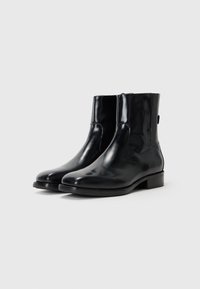 Botas de tobillo de charol negro con un acabado suave, punta redondeada y tacón bajo cuadrado; cuentan con paneles elásticos laterales y una sutil lengüeta trasera.