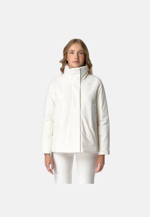 Donna che indossa una giacca a vento bianca con collo alto e pantaloni bianchi, in piedi davanti a uno sfondo grigio chiaro uniforme.