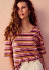 Gestreiftes Kurzarmoberteil mit lila, orangefarbenen und weißen Farbtönen; weicher Stoff mit lockerem Schnitt; Rundhalsausschnitt; getragen zu hellen Hosen.