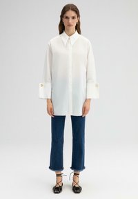 Witte blouse met lange mouwen en een puntige kraag, met grote manchetten versierd met gouden knopen. Gecombineerd met blauwe jeans met rafels aan de onderkant en zwarte schoenen.