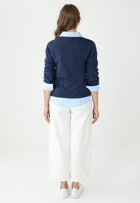 Granatowy sweter z prążkowanym wykończeniem noszony na jasnoniebieską koszulę w paski, zestawiony z białymi szerokimi spodniami i białymi trampkami, widziany od tyłu.