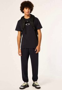 Ungdom med krøllete hår iført svart t-skjorte, svarte bukser, svart ryggsekk og svart-hvite sneakers står mot en ensfarget bakgrunn.