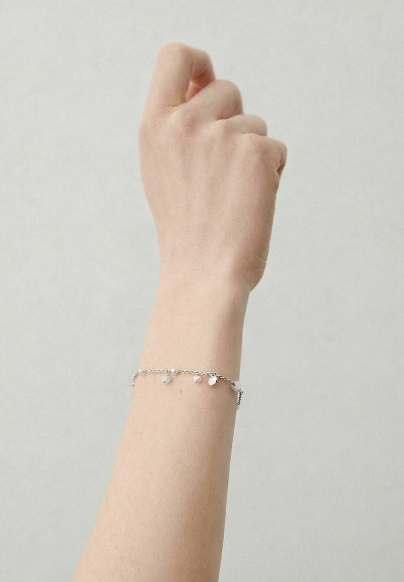 Main gauche à la peau claire, doigts recourbés vers l'intérieur, portant un délicat bracelet en argent avec de petits charms ronds, sur un fond uni.