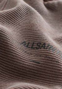 Teksturēts audums maiga rozā krāsā, ar ribotu rakstu un tumši pelēkā krāsā iespiestu tekstu "ALLSAINTS". Mīksts, plūstošs materiāls.