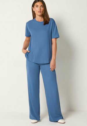 Une femme porte un haut bleu à manches courtes et un pantalon à jambes larges. Elle se tient debout, une main dans la poche, devant un fond de studio uni.