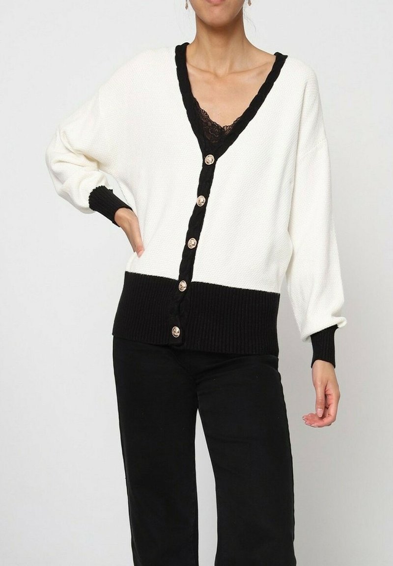 Cardigan en tricot gaufré blanc avec un col en V noir et des poignets noirs. Présente des accents de boutons noirs et une coupe ample. Le tissu texturé ajoute du détail.