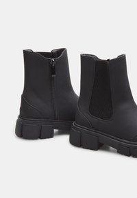 Botas negras hasta el tobillo con un acabado suave y mate, que cuentan con una cremallera lateral y paneles elásticos, y una suela de goma texturizada y gruesa.