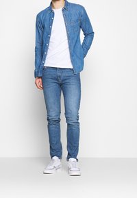 Denimskjorta i ljusblått med knappar och bröstficka, matchad med medelblå jeans och vita sneakers. Totalt avslappnad outfit.