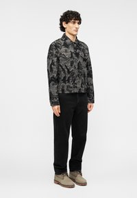 Μαύρο ανδρικό τζιν μπουφάν με σχέδια και δύο τσέπες στο στήθος, συνδυασμένο με μαύρο παντελόνι και γκρι παπούτσια με κορδόνια, σε ουδέτερο φόντο.