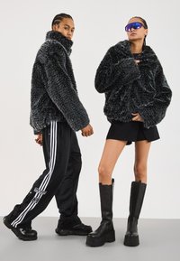 Zwei Modelle tragen schwarze und graue, strukturierte Kunstfelljacken; eines in schwarzen Adidas Jogginghosen und Sneakern, das andere in schwarzen Stiefeln und Shorts.