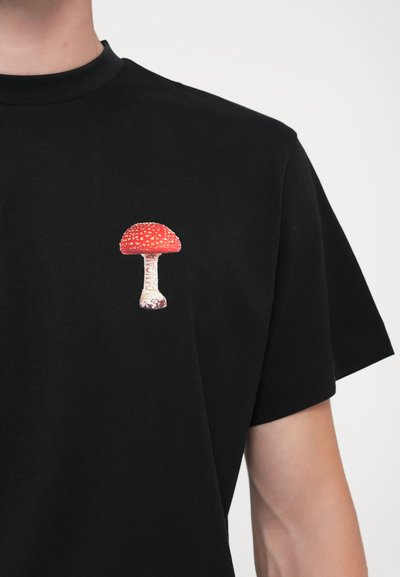 T-shirt noir à manches courtes avec un motif de champignon rouge et blanc sur la partie gauche de la poitrine. Texture de tissu lisse.