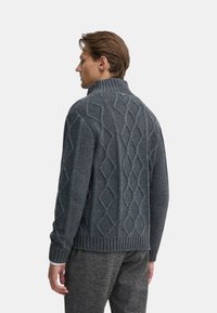 Maglione grigio a maglia con colletto alto, caratterizzato da un motivo a diamante testurizzato e orlo a costine. Indossato sopra pantaloni grigi a fantasia.