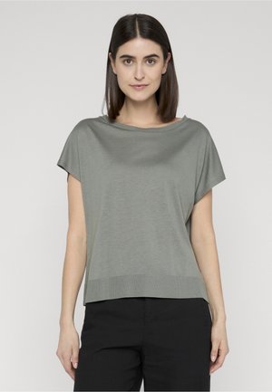 Kvinne med rett mørkt hår som har på seg en løs grå kortermet topp og svarte bukser, stående mot en ensfarget bakgrunn.